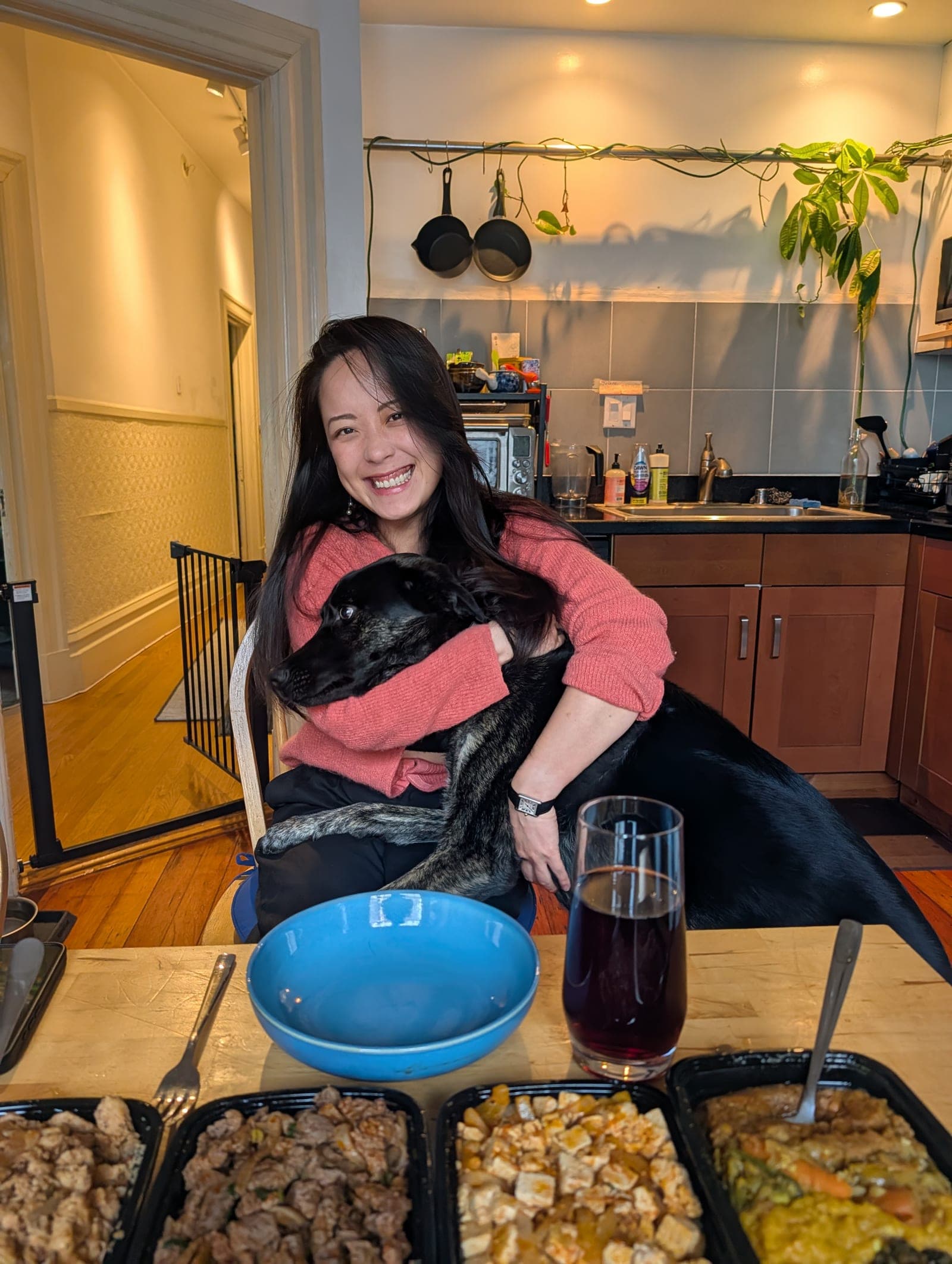 Cecilia in a coral sweater laughing as she holds her black dog at a kitchen table, takeout containers and a glass of red wine in front of them.