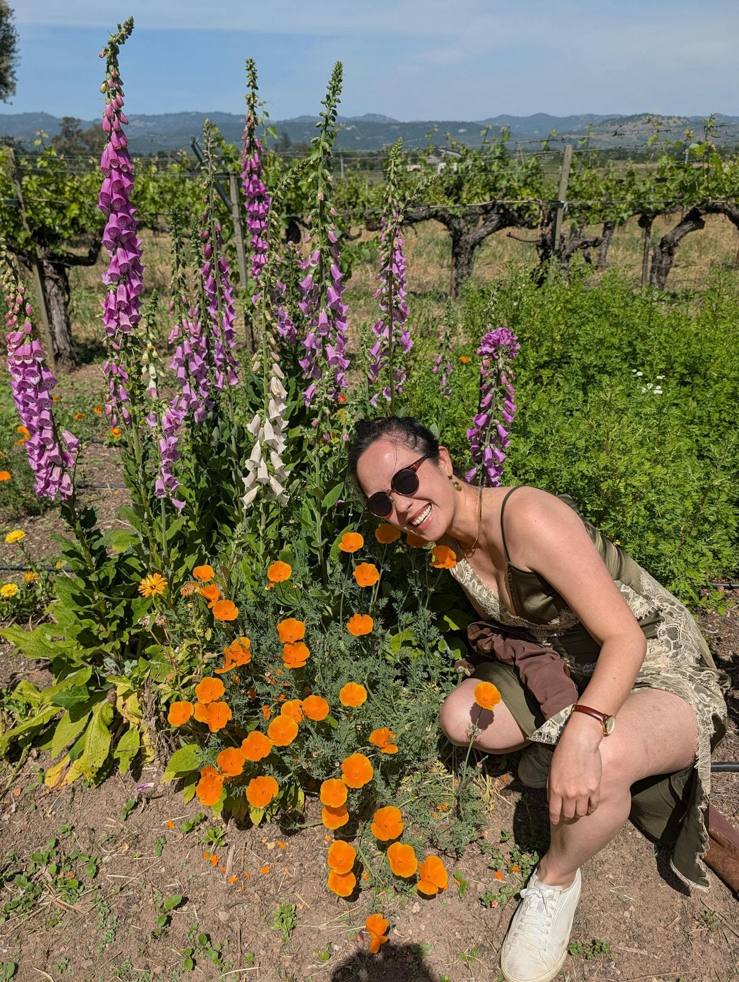 Cecilia crouched beside a bed of California poppies and tall pink foxgloves, sunglasses on, vineyard rows and hills behind her on a sunny day.
